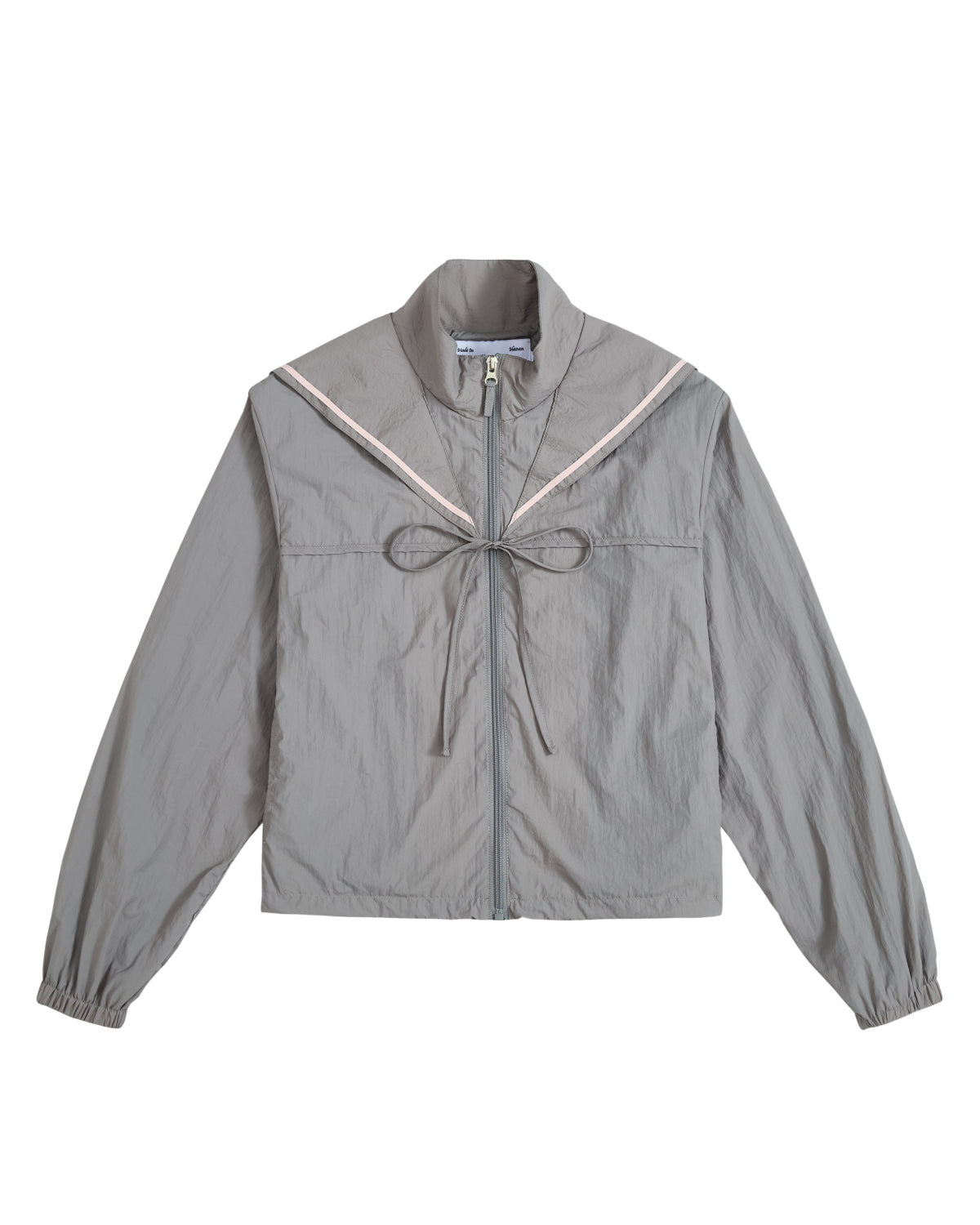 The GREY SAILOR WINDBREAKER by Sundae School is a light gray cropped jacket featuring a high collar, long sleeves, and a decorative tie at the chest. A subtle pink chevron detail on the front evokes the look of a sailor uniform, while the slightly crinkled fabric adds both style and comfort.