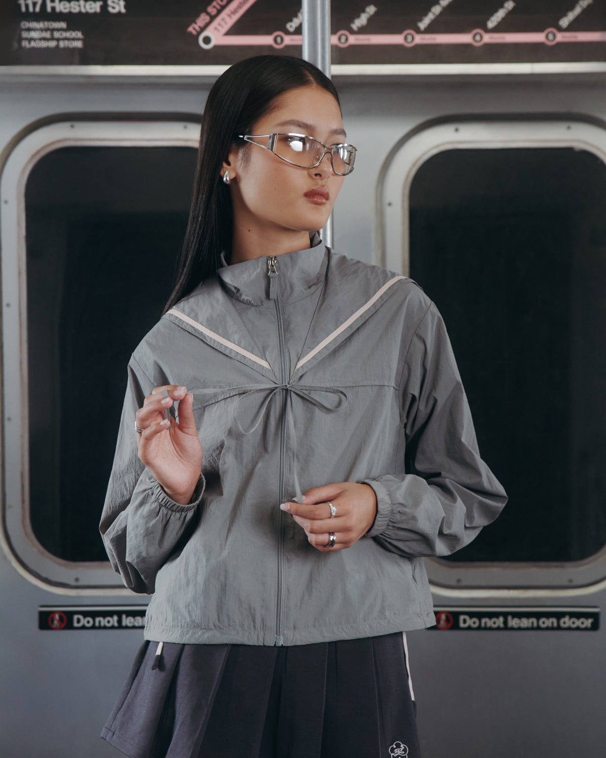 A woman with long dark hair and glasses stands by subway doors, wearing the Sundae School CHARCOAL SAILOR WINDBREAKER and a dark skirt. She looks to the side, holding her hair, with a subway map in the background.