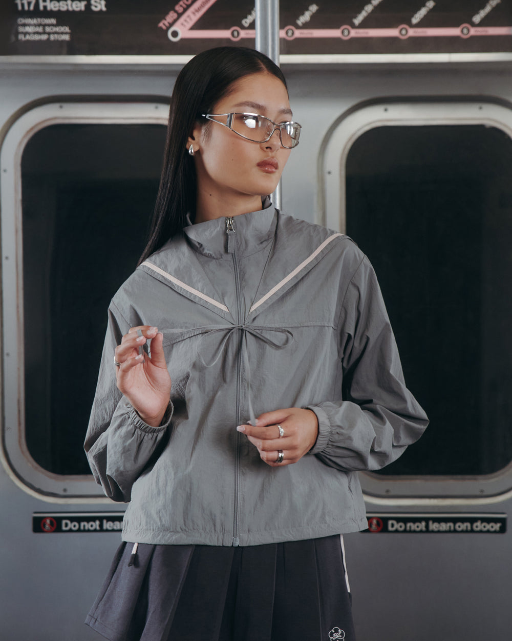A woman in glasses and the Sundae School GREY SAILOR WINDBREAKER stands in a subway car, holding a pen. Behind her are subway doors and a blurred map as she looks to the side with a calm expression.