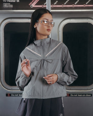 A woman with long dark hair and glasses stands by subway doors, wearing the Sundae School CHARCOAL SAILOR WINDBREAKER and a dark skirt. She looks to the side, holding her hair, with a subway map in the background.