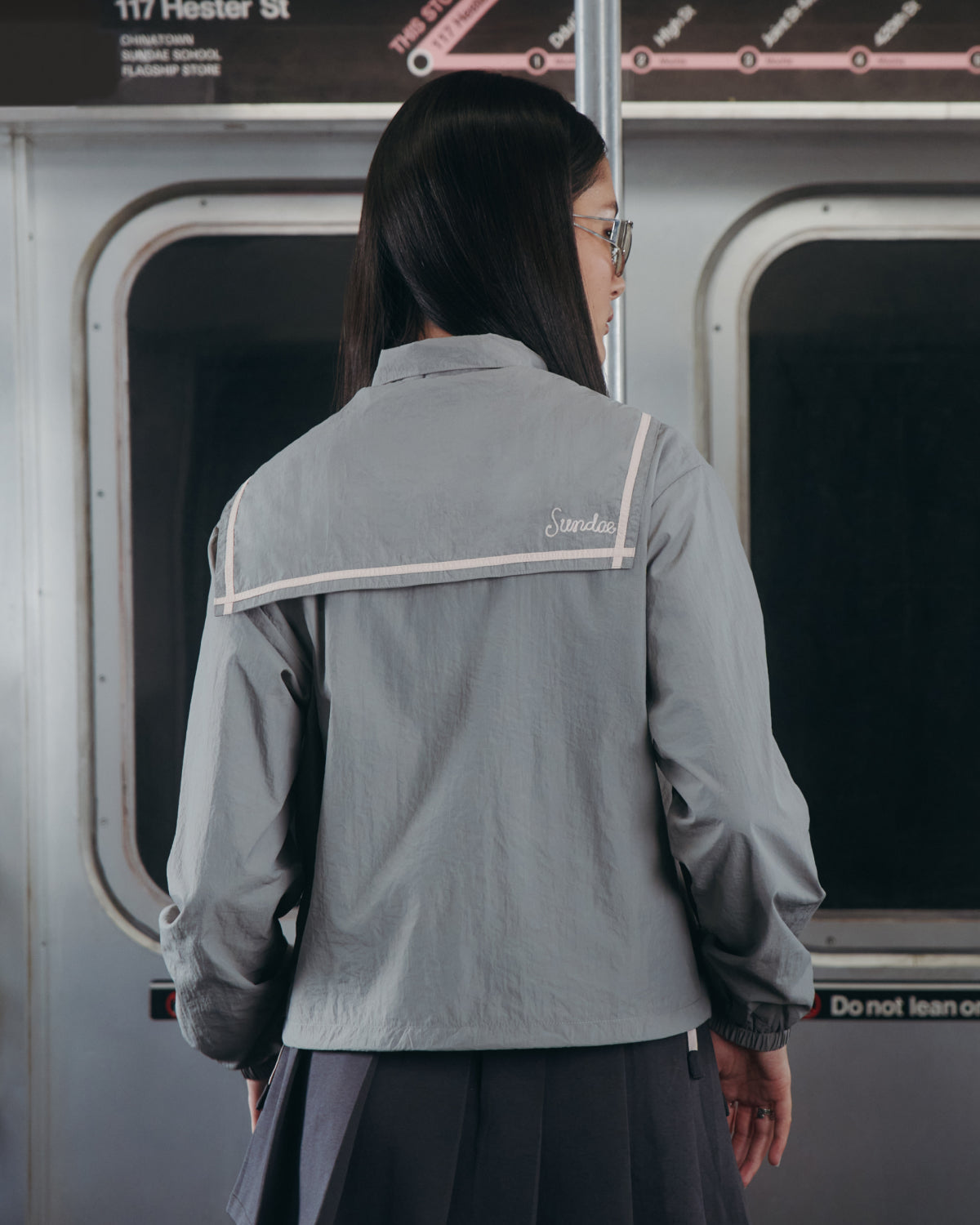 A person with long dark hair and glasses, wearing the Sundae School CHARCOAL SAILOR WINDBREAKER, stands facing away in a subway train. Subway station signs and sliding doors appear in the background.