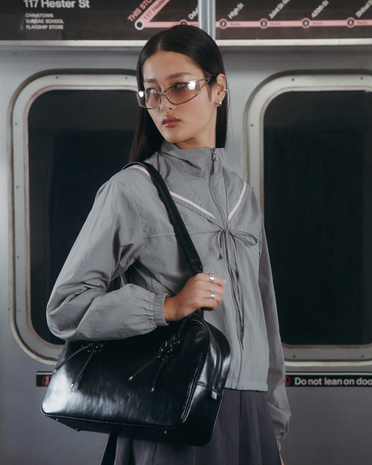 A woman in clear glasses and the CHARCOAL SAILOR WINDBREAKER by Sundae School stands on a subway train, holding a large black handbag. Subway windows and a station map are visible in the background.