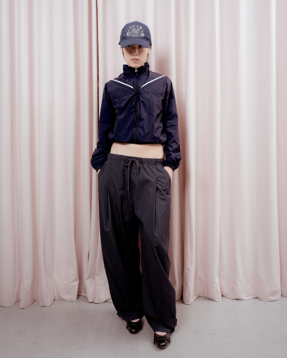 Full-length front view of a person with short black hair standing against a light pink curtain backdrop. They are wearing a dark navy baseball cap, a cropped navy windbreaker jacket with white piping details at the shoulders, and wide-leg dark pinstripe trousers with their hands in their pockets.