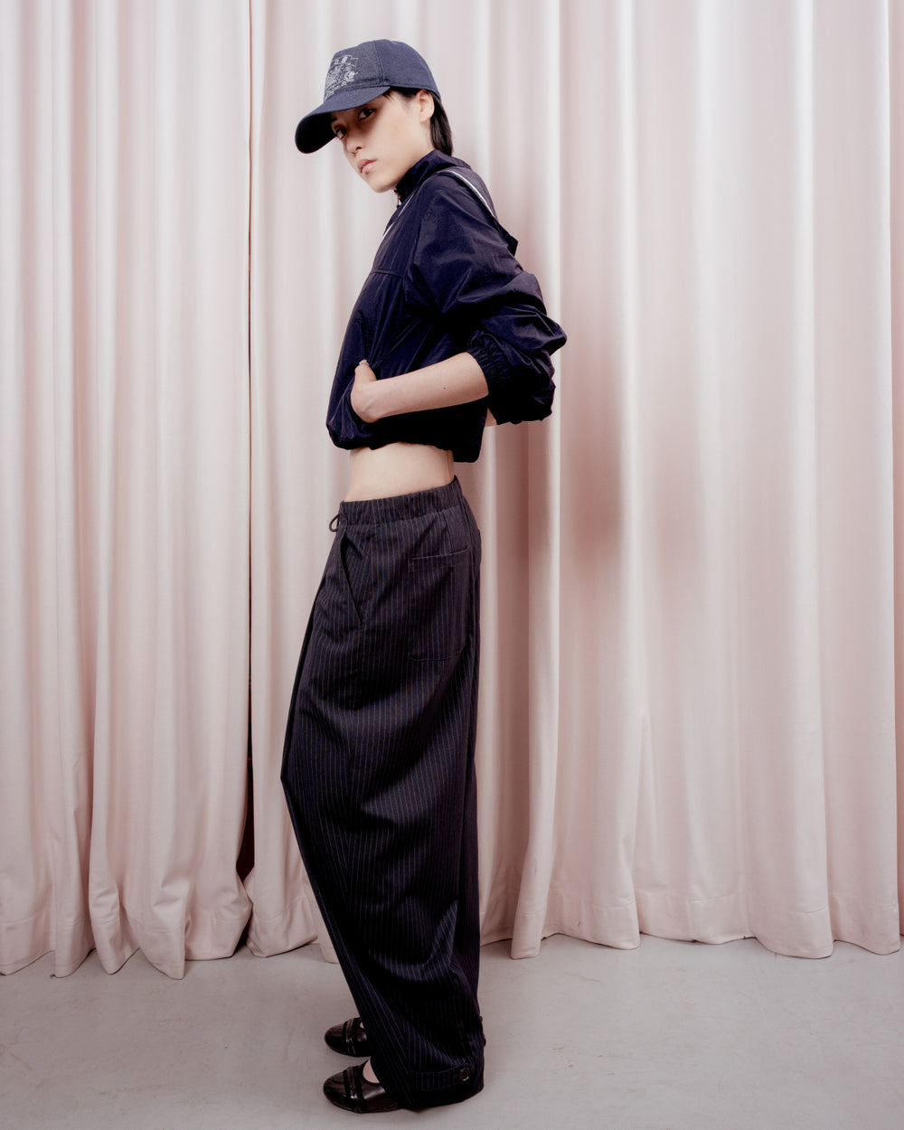 Full-length side profile view of a person with short black hair looking toward the camera against a pink curtain backdrop. They are wearing a dark navy baseball cap, a cropped navy windbreaker jacket, wide-leg dark pinstripe trousers, and black shoes.