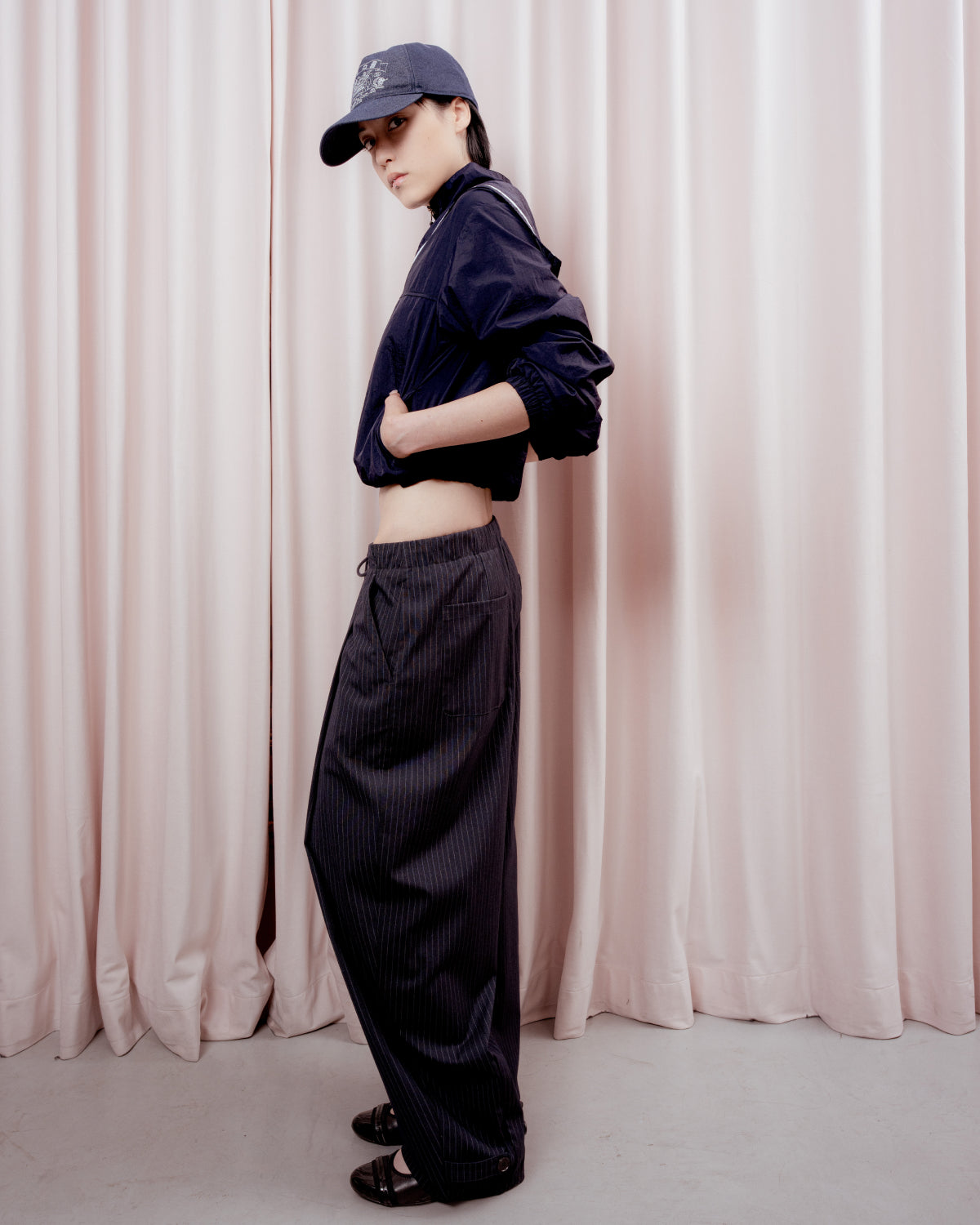 Full-length side profile view of a person with short black hair looking toward the camera against a pink curtain backdrop. They are wearing a dark navy baseball cap, a cropped navy windbreaker jacket, wide-leg dark pinstripe trousers, and black shoes.