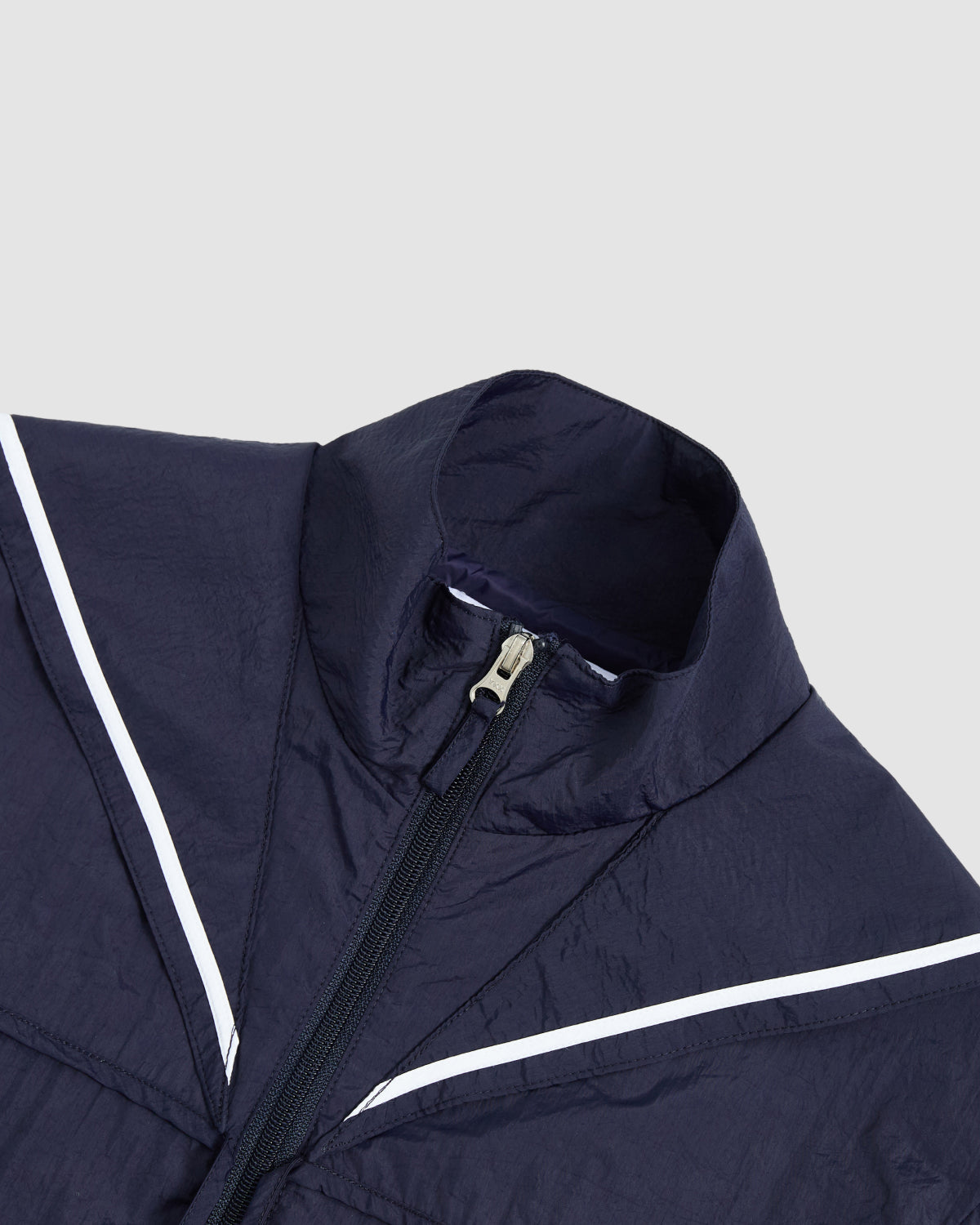 The NAVY SAILOR WINDBREAKER by Sundae School features a high collar, front zipper, and white diagonal stripes across the chest and shoulders, shown here in a close-up on a light grey background.