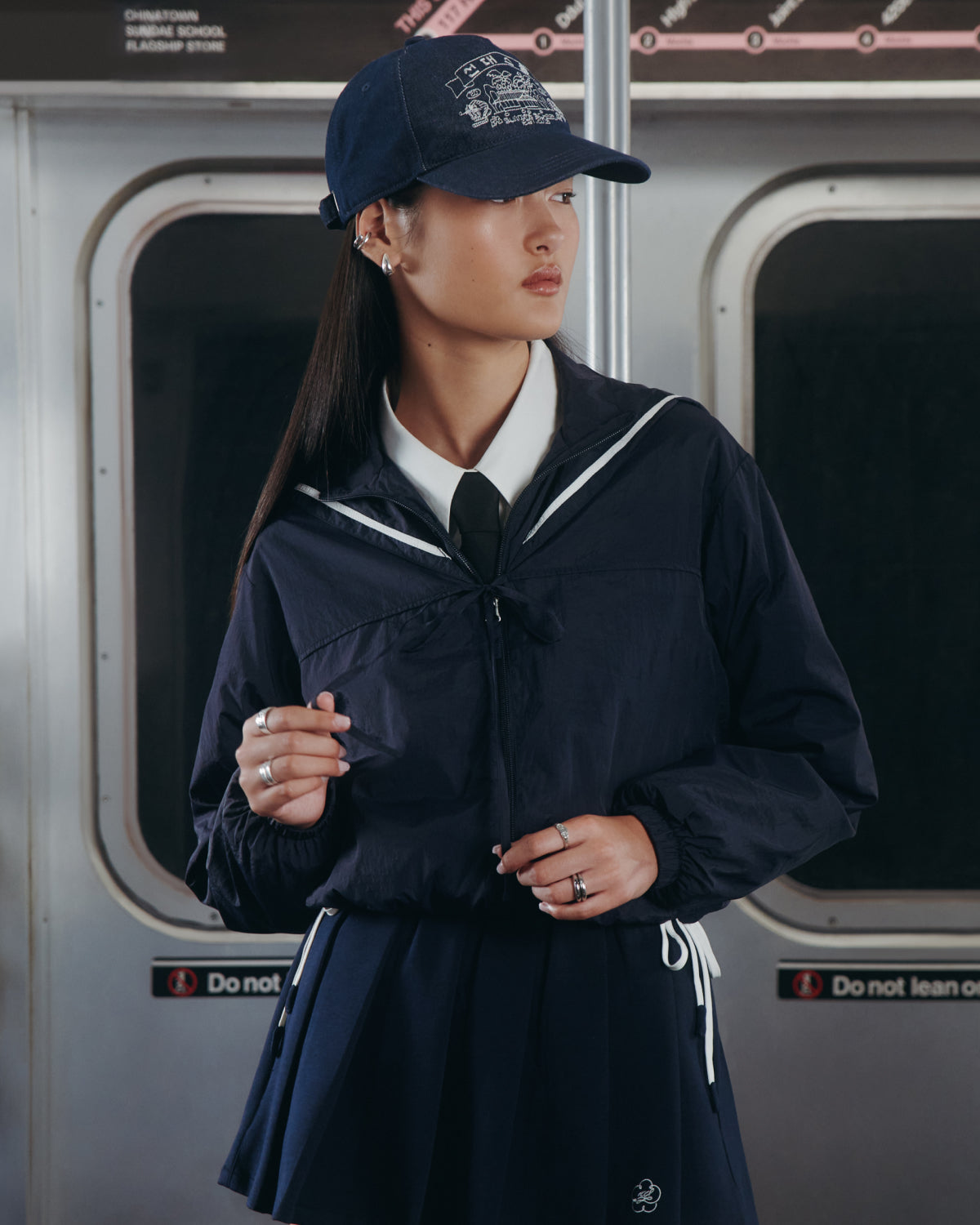 A person in Sundae School's NAVY SAILOR WINDBREAKER stands in a subway car, pairing the jacket with a matching skirt, white shirt, black tie, and a UV-protective navy baseball cap, as subway doors and a route map appear behind them.