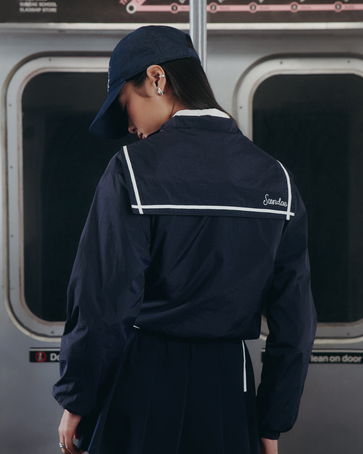 A person in a Sundae School NAVY SAILOR WINDBREAKER and cap stands with their back to the camera on a subway train, "Standard" written in white on the jacket's collar.