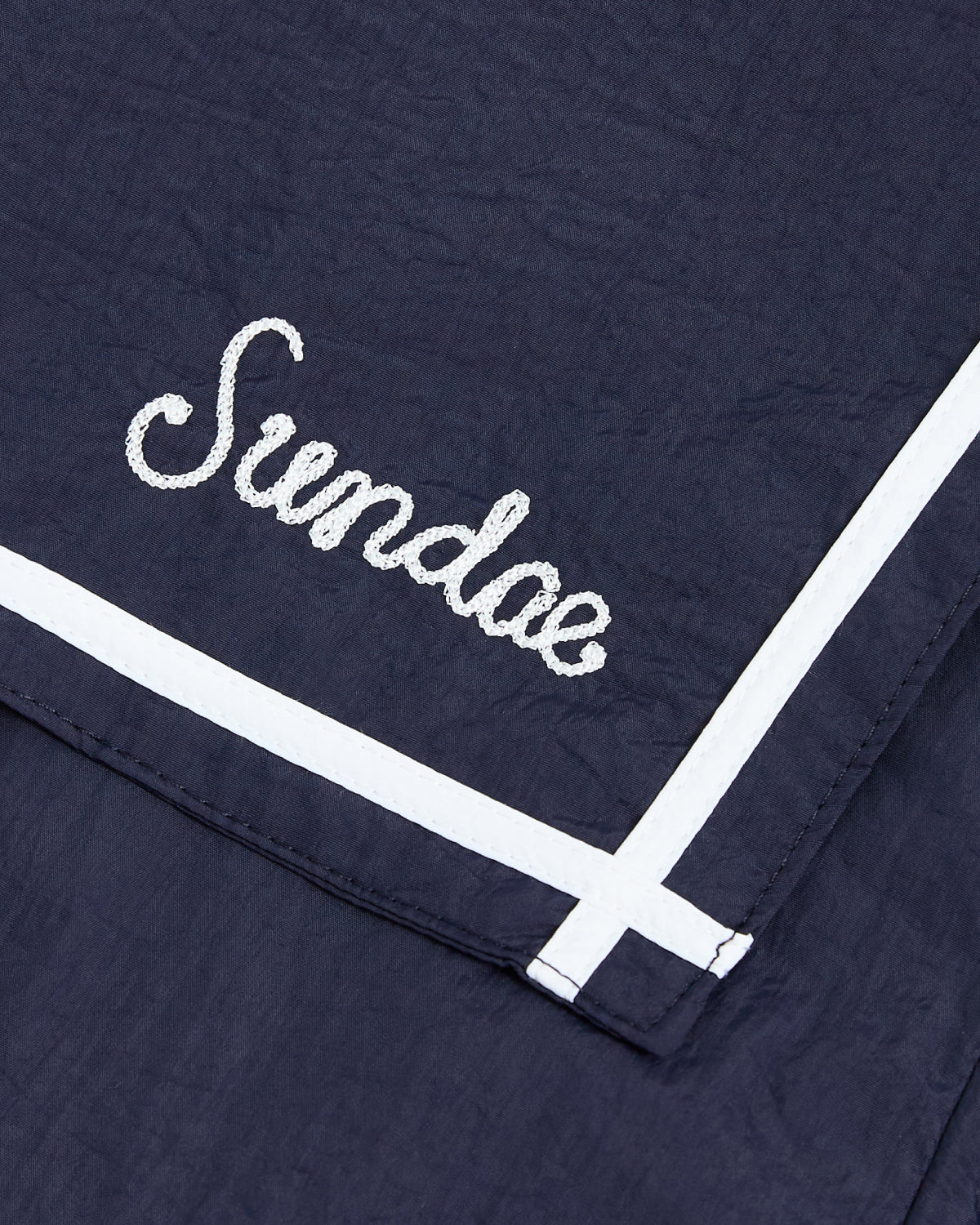 Crafted from navy blue fabric, the NAVY SAILOR WINDBREAKER by Sundae School features “Sundae” embroidered in crisp white cursive. Two bold white stripes accentuate one corner, reminiscent of classic sailor uniform jacket designs.