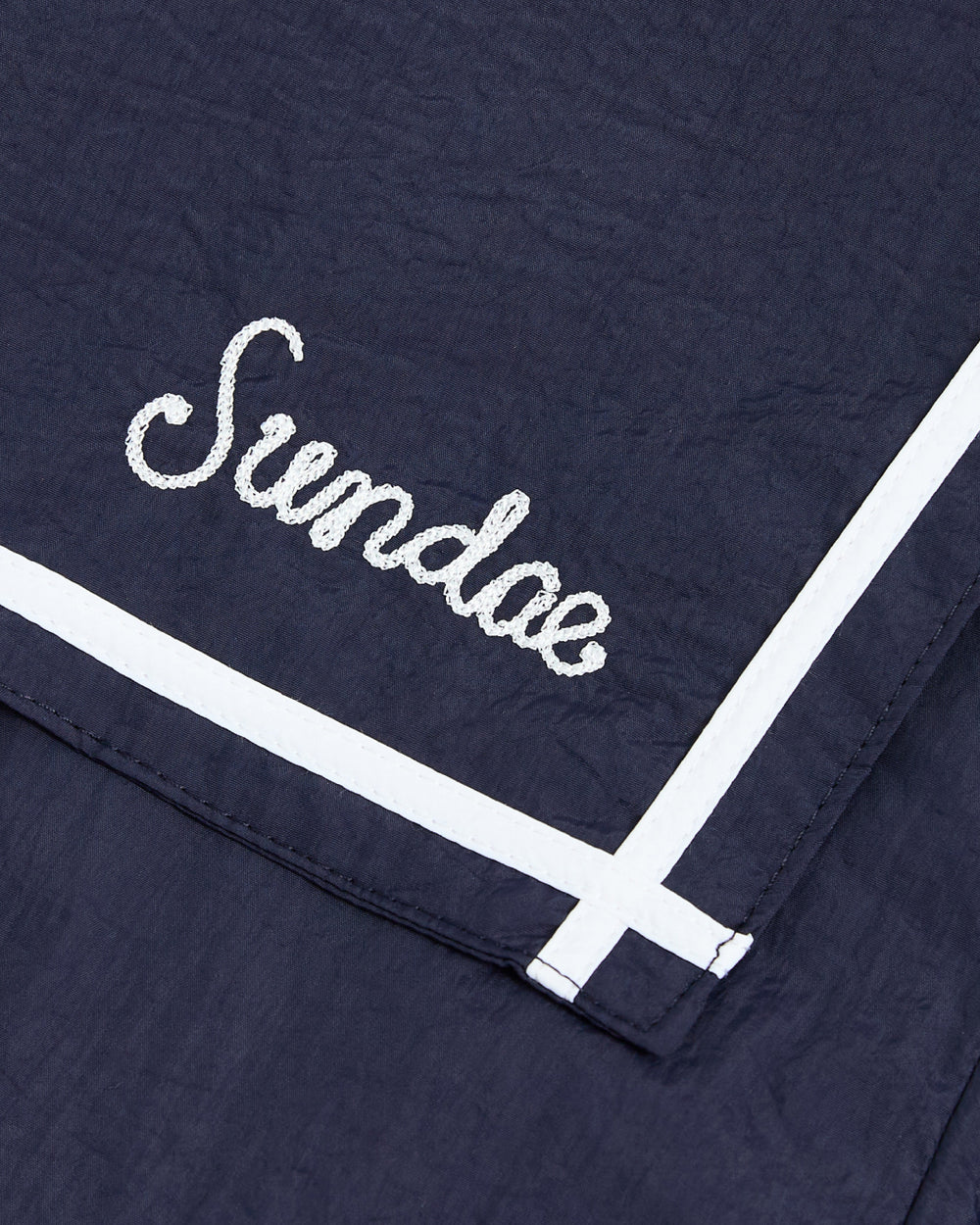 Crafted from navy blue fabric, the NAVY SAILOR WINDBREAKER by Sundae School features “Sundae” embroidered in crisp white cursive. Two bold white stripes accentuate one corner, reminiscent of classic sailor uniform jacket designs.