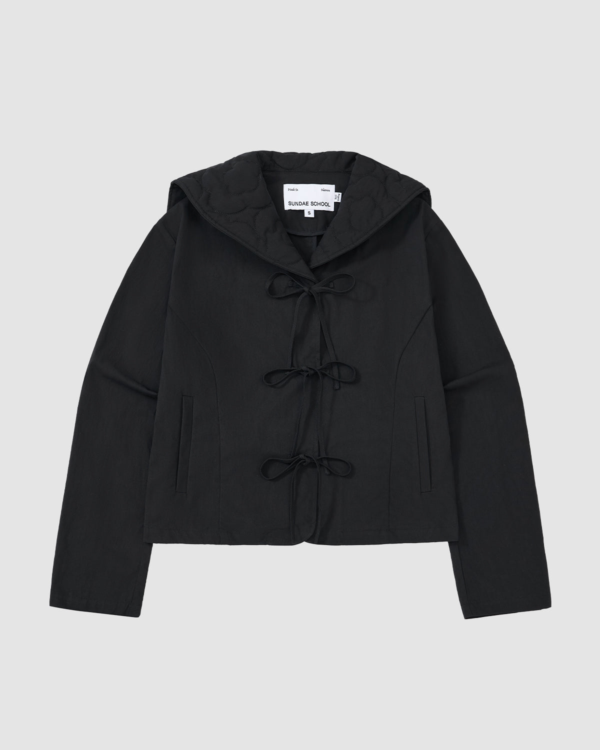 The BLACK SAILOR COLLAR UNIFORM JACKET by Sundae School is a black hooded jacket with mochi quilted stitching, three bow tie closures, and two vertical slit pockets. The inside collar features a visible label.