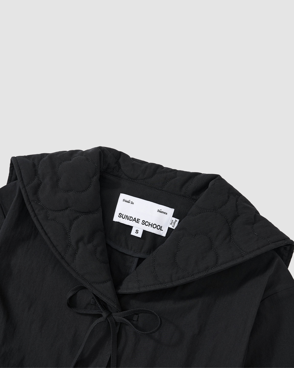 A detailed view of the BLACK SAILOR COLLAR UNIFORM JACKET by Sundae School shows mochi quilted stitching, a floral pattern, and a label with “Sundae School” and size S, evoking a Korean school uniform style. Additional tags are partially visible.