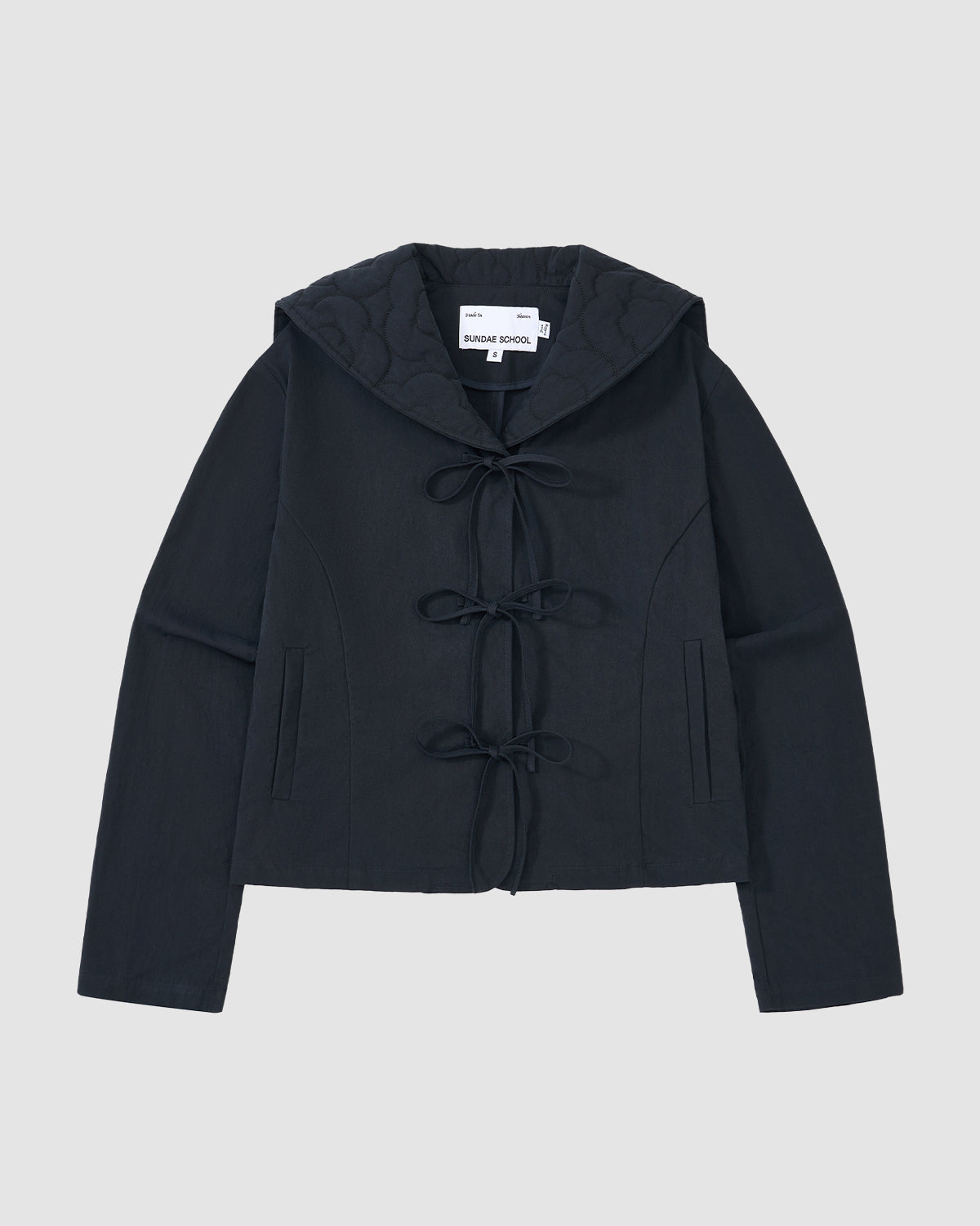 The NAVY SAILOR COLLAR UNIFORM JACKET by Sundae School features a dark navy hue, mochi quilted stitching, long sleeves, hood, front tie closures, two vertical pockets, and an inner collar label, shown on a light gray background.
