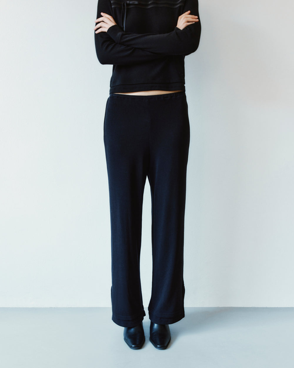 A person with arms crossed wears a black long-sleeved top and BLACK SEONGSU APRON PANTS, which are black wide-leg trousers with an adjustable waistband.