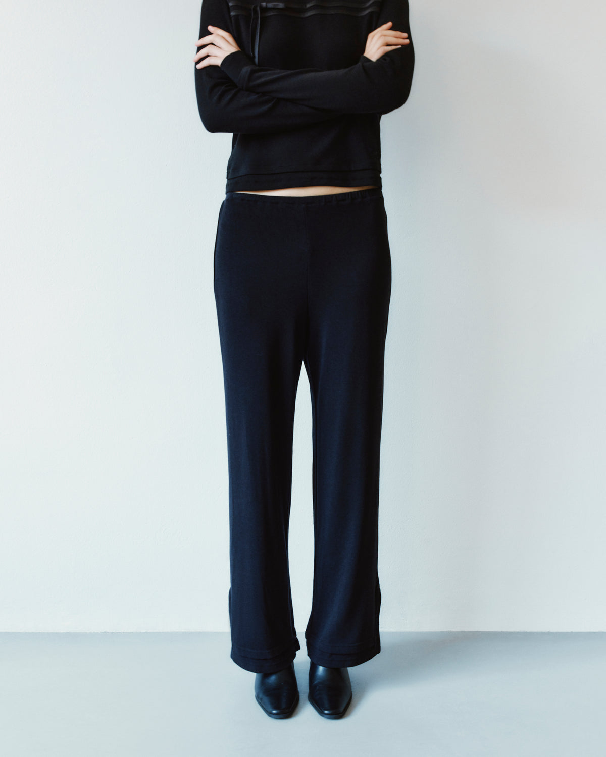 A person with arms crossed wears a black long-sleeved top and BLACK SEONGSU APRON PANTS, which are black wide-leg trousers with an adjustable waistband.