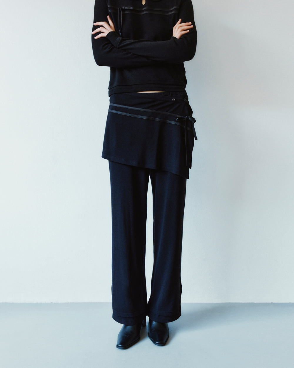 A person wears a black long-sleeved shirt with arms crossed, paired with black wide-leg BLACK SEONGSU APRON PANTS featuring a hybrid wrap skirt overlay.
