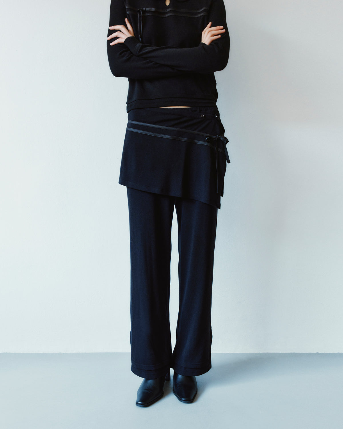A person wears a black long-sleeved shirt with arms crossed, paired with black wide-leg BLACK SEONGSU APRON PANTS featuring a hybrid wrap skirt overlay.