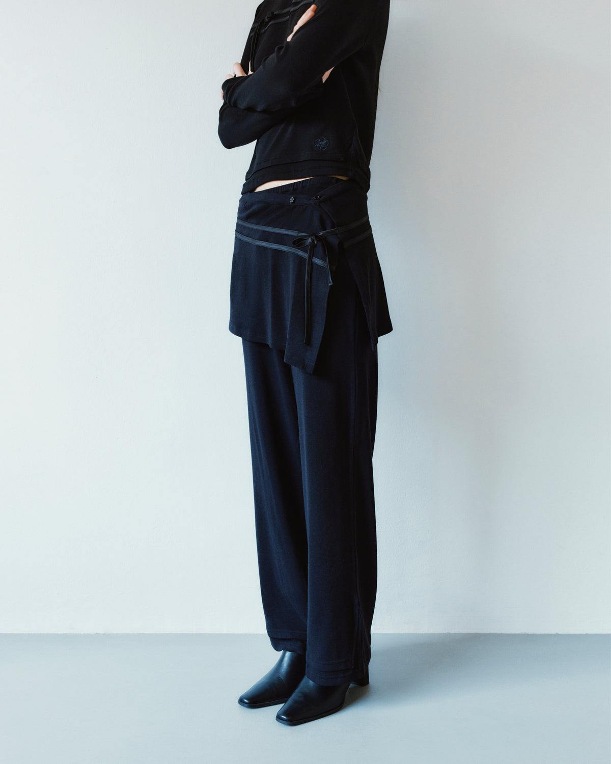 A person in profile wears black Sundae School BLACK SEONGSU APRON PANTS with an integrated hybrid wrap skirt panel at the waist, finished with black leather boots.