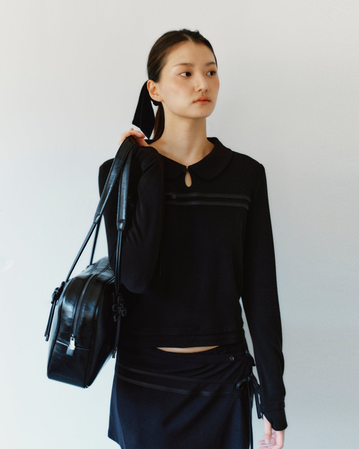 A person wearing a black rounded collar BLACK SEONGSU KEYHOLE TOP with a small keyhole cutout at the neck, featuring satin ties and carrying a black shoulder bag.