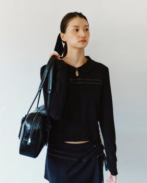 A person wearing a black rounded collar BLACK SEONGSU KEYHOLE TOP with a small keyhole cutout at the neck, featuring satin ties and carrying a black shoulder bag.