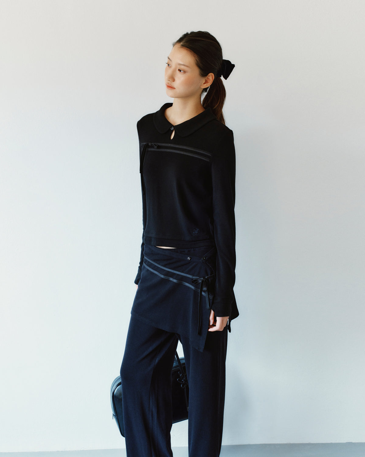 A person wears a black Sundae School BLACK SEONGSU KEYHOLE TOP with a rounded collar and satin ties, paired with matching black trousers that feature a wrap-style skirt overlay.