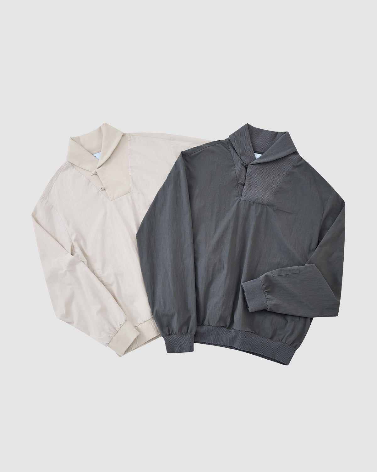Two IVORY SHAWL COLLAR ANORAKS by Sundae School—one beige, one dark gray—with shawl collars, elastic cuffs and hem, and cotton-nylon shells, are laid flat side by side on a plain white background.
