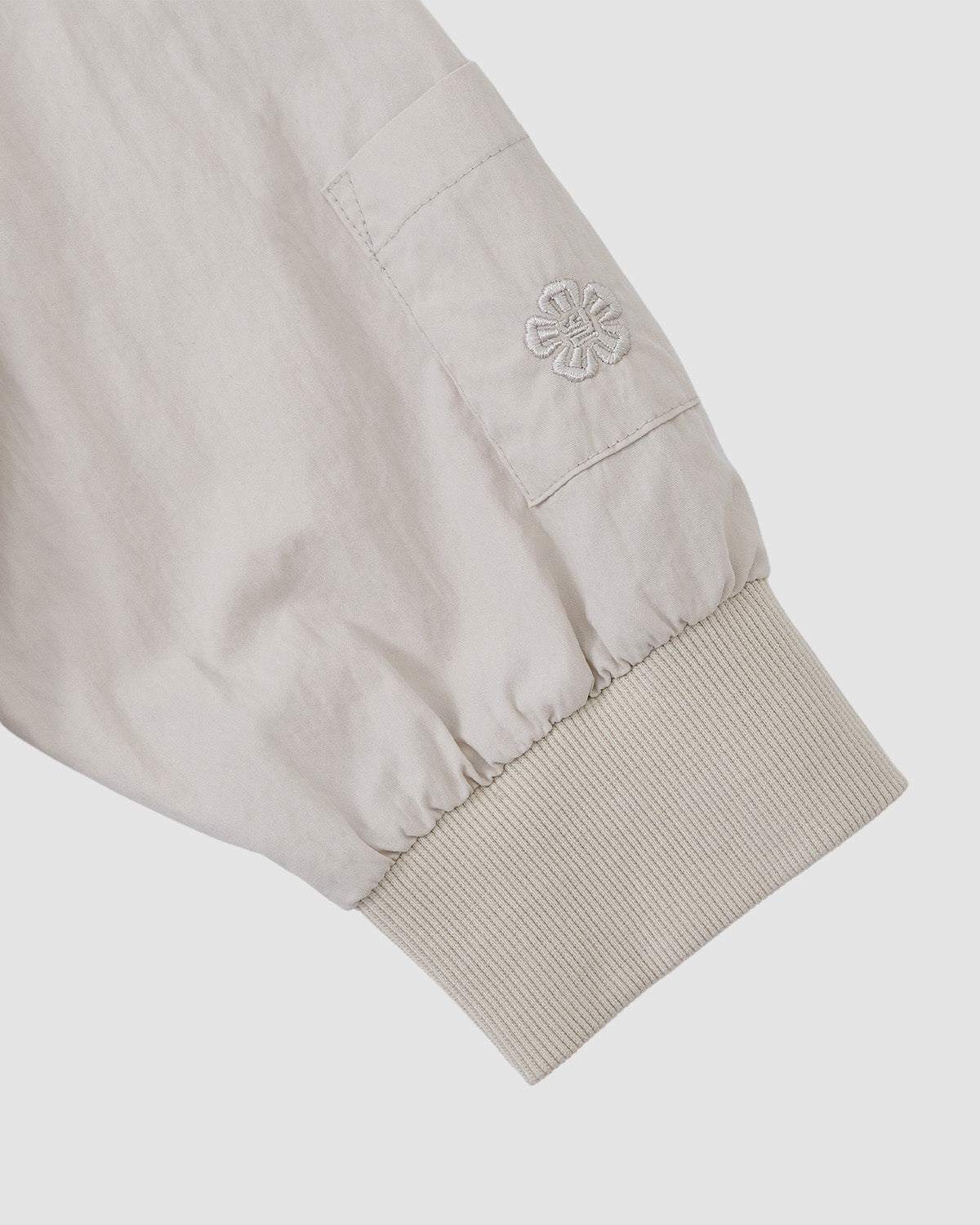 A close-up of the Sundae School IVORY SHAWL COLLAR ANORAK shows its light beige sleeve with a ribbed knit cuff, small pocket, and embroidered flower above the pocket. The cotton-nylon shell is set against a plain white background.