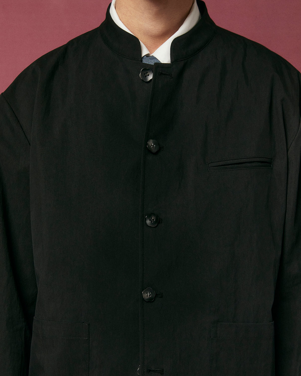 A person models the BLACK STAND COLLAR UNIFORM JACKET by Sundae School over a white shirt, shown from shoulders to mid-torso against a muted red background. Their face is not visible.