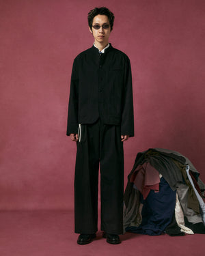 A person in Sundae School BLACK PLEATED UNIFORM SLACKS and black sunglasses stands before a pink backdrop, beside a pile of assorted clothes stacked on the floor.