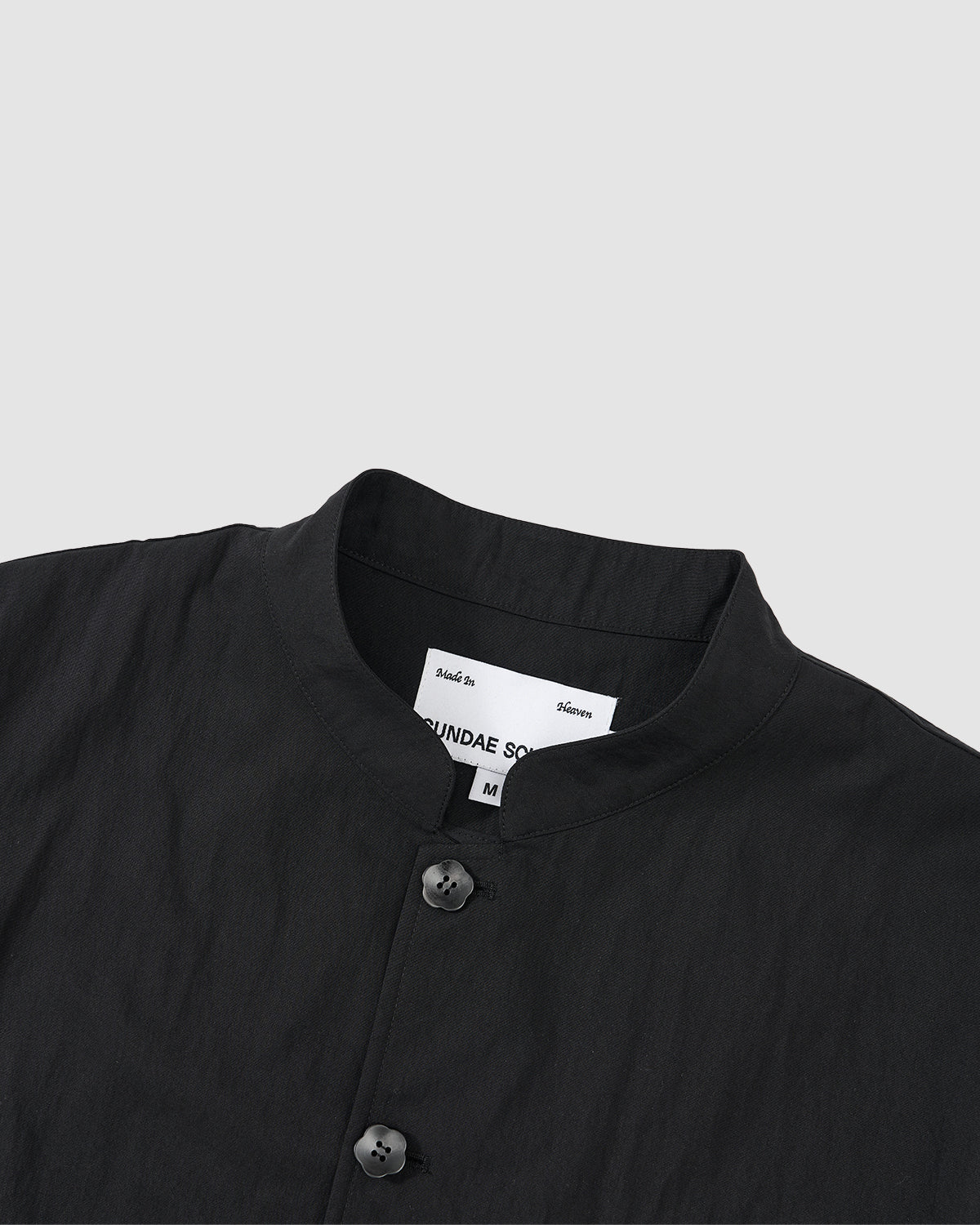 Close-up of the collar and upper chest of the BLACK STAND COLLAR UNIFORM JACKET by Sundae School, crafted from a cotton nylon blend. Features a stand collar, two black buttons, and an inner white “SUNDAE SCHOOL” label.