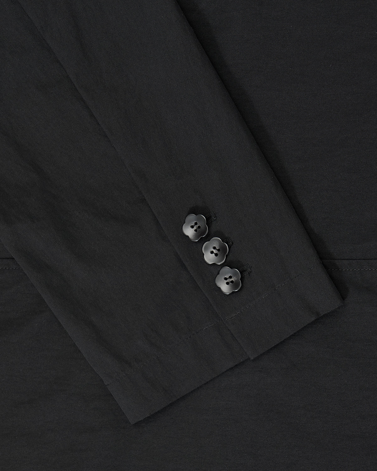 Close-up of the BLACK STAND COLLAR UNIFORM JACKET by Sundae School, showing its black sleeve with three black buttons near the cuff, crafted from a cotton nylon blend and set against matching fabric.