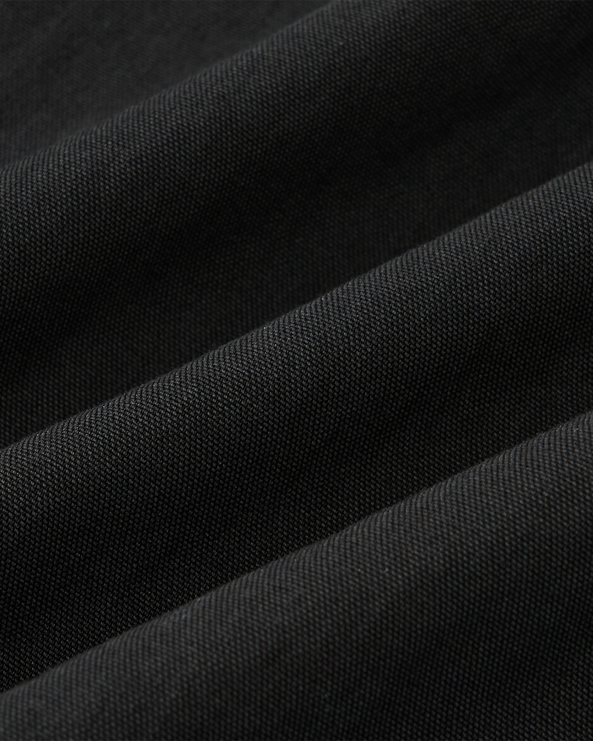 Close-up of the BLACK STAND COLLAR UNIFORM JACKET by Sundae School, featuring black cotton nylon blend fabric with diagonal folds and a fine textured weave—highlighted by lighting to show the material’s subtle ridges and pattern.