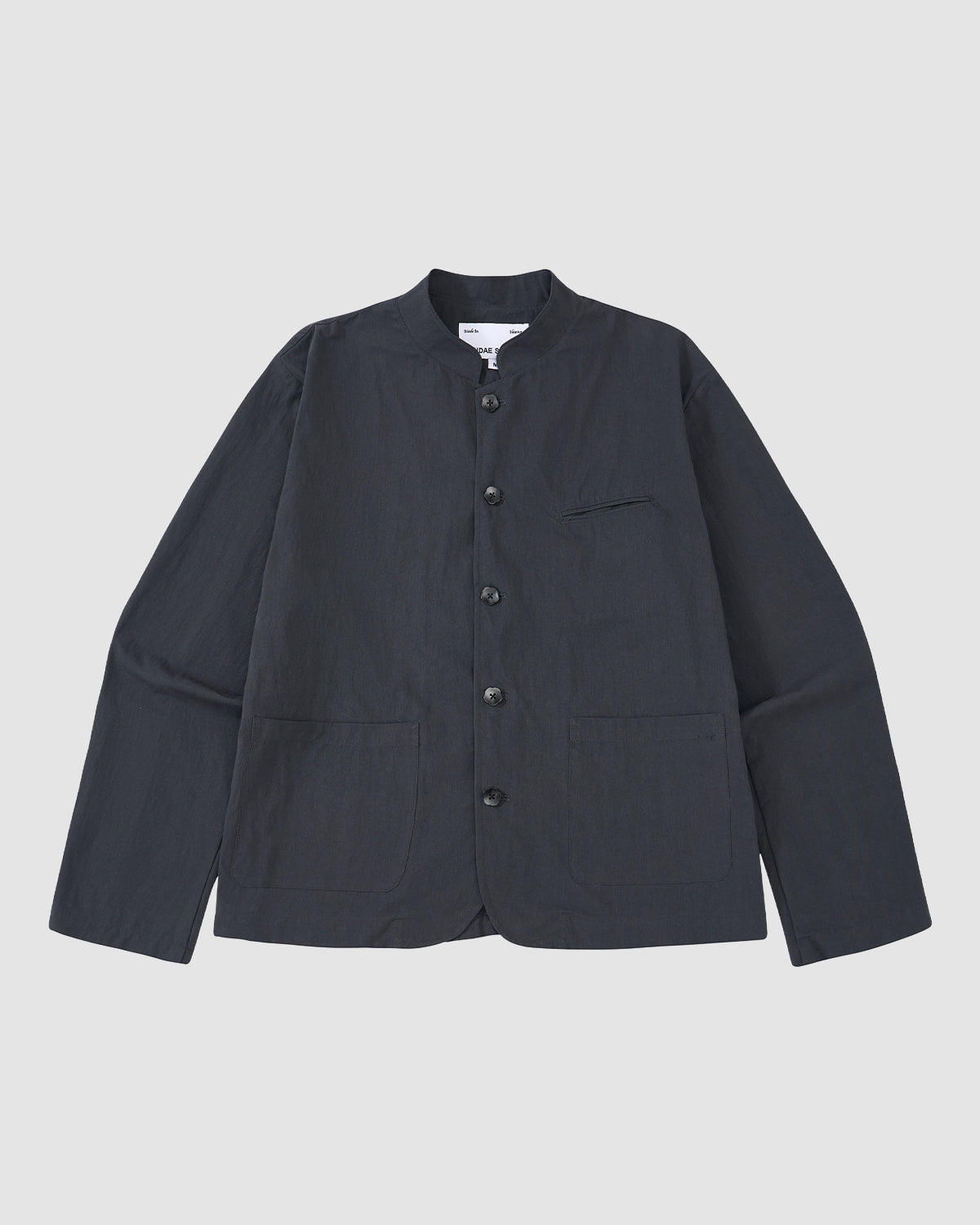 The Sundae School CHARCOAL STAND COLLAR UNIFORM JACKET, made from a cotton-nylon blend with long sleeves and three front pockets, is shown laid flat on a light gray background.