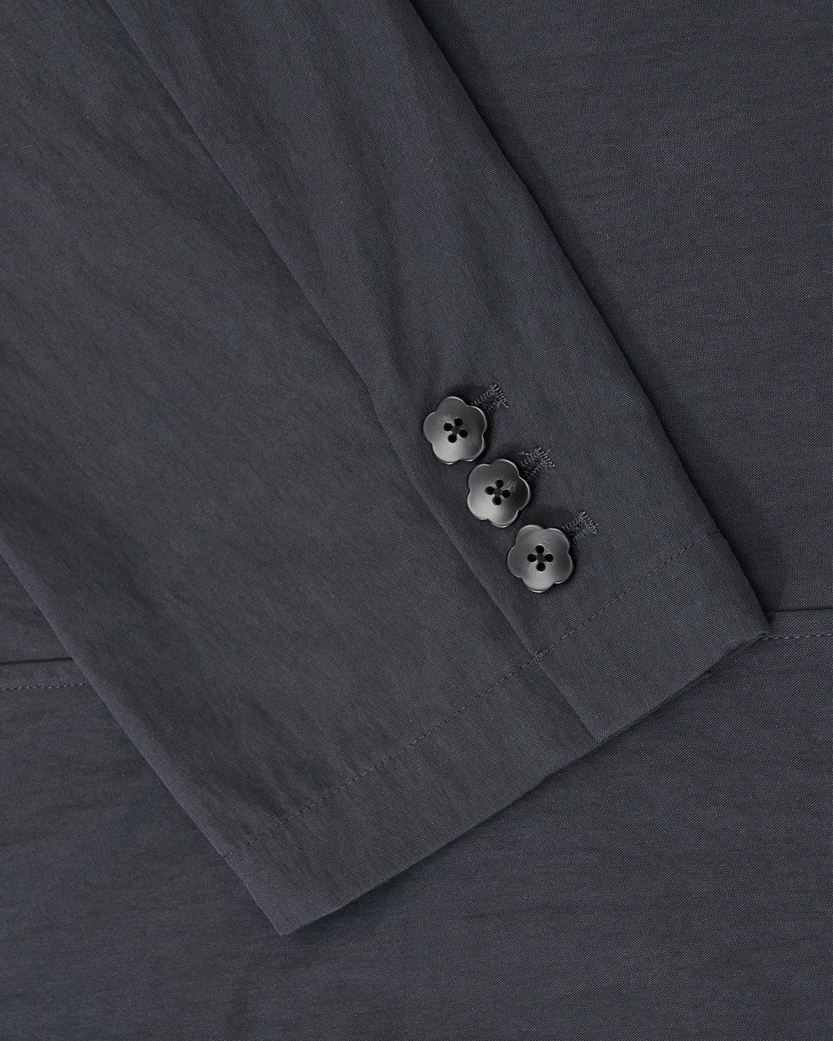 A close-up of the CHARCOAL STAND COLLAR UNIFORM JACKET by Sundae School shows its sleeve with three black flower-shaped buttons on the cuff, crafted from a dark gray cotton nylon blend.
