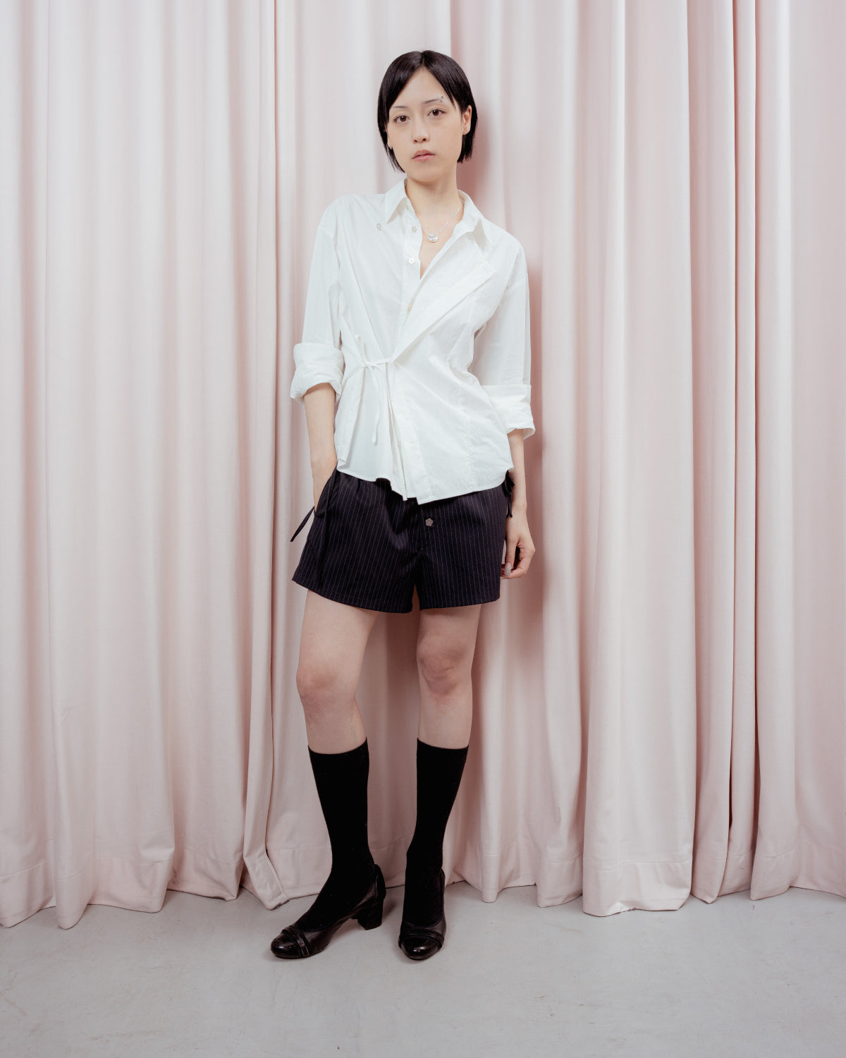 A person with short dark hair stands in front of light pink curtains, wearing a white button-up shirt, Sundae School STRIPED BOXER SHORTS, long black socks, and black shoes.