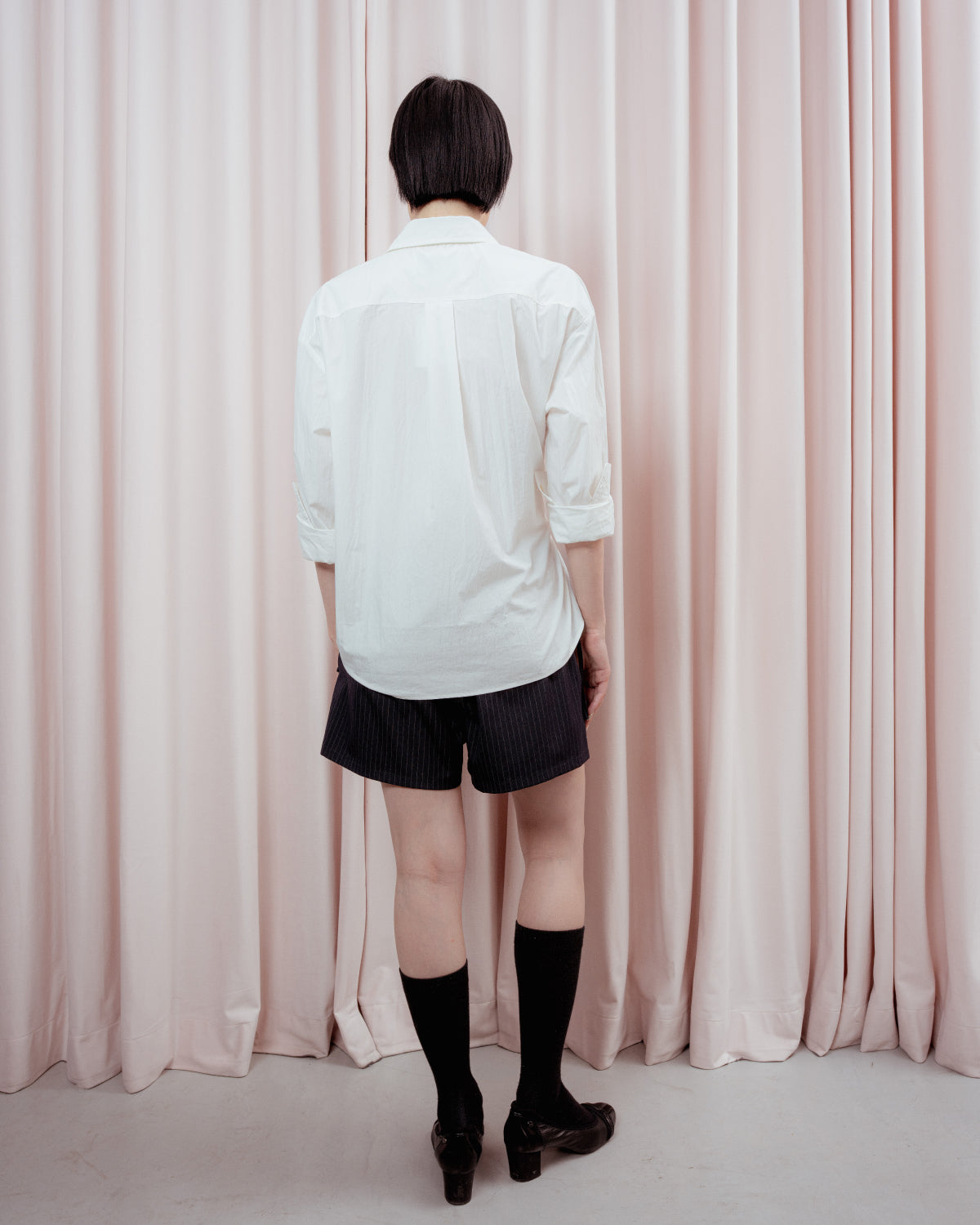 A person with short dark hair, wearing a white shirt and Sundae School STRIPED BOXER SHORTS, knee-high black socks, and black shoes, stands facing light pink curtains with their back to the camera.