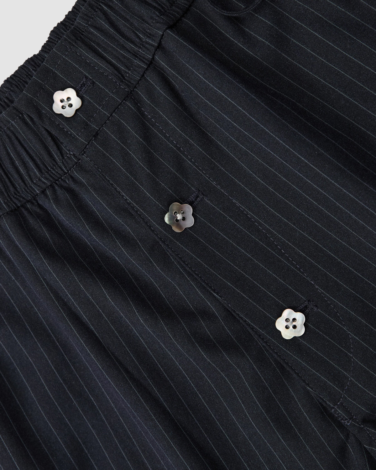 Close-up of black pinstripe fabric with white flower-shaped buttons and an elastic waistband, featured on the STRIPED BOXER SHORTS by Sundae School.
