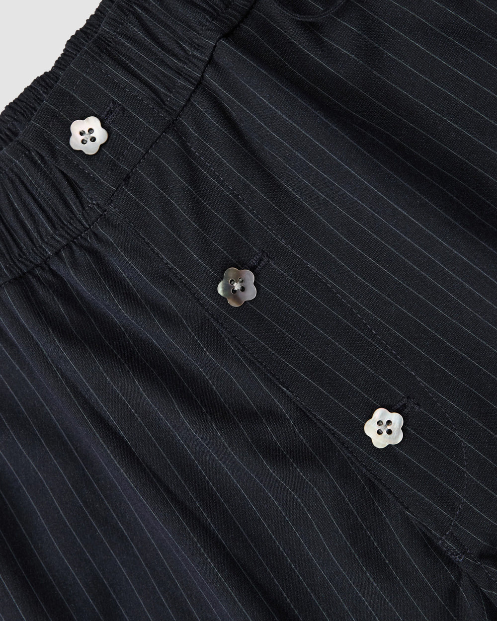 Close-up of black pinstripe fabric with white flower-shaped buttons and an elastic waistband, featured on the STRIPED BOXER SHORTS by Sundae School.