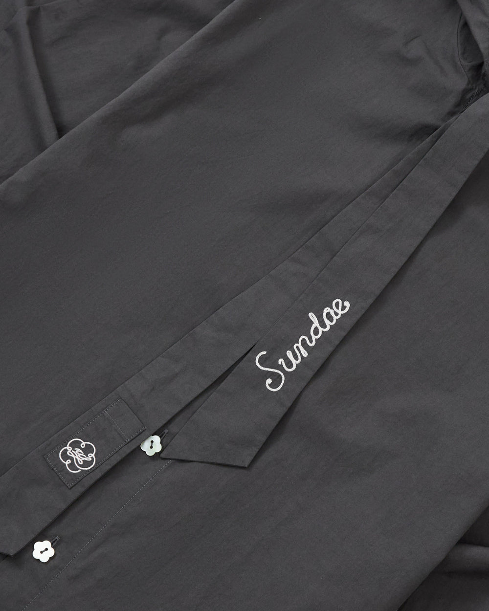 A close-up of the CHARCOAL SUNBAE TIE SHIRT by Sundae School shows white embroidery—one area spells out “Sundae,” while another features a small flower-shaped button topped with a cloud design.