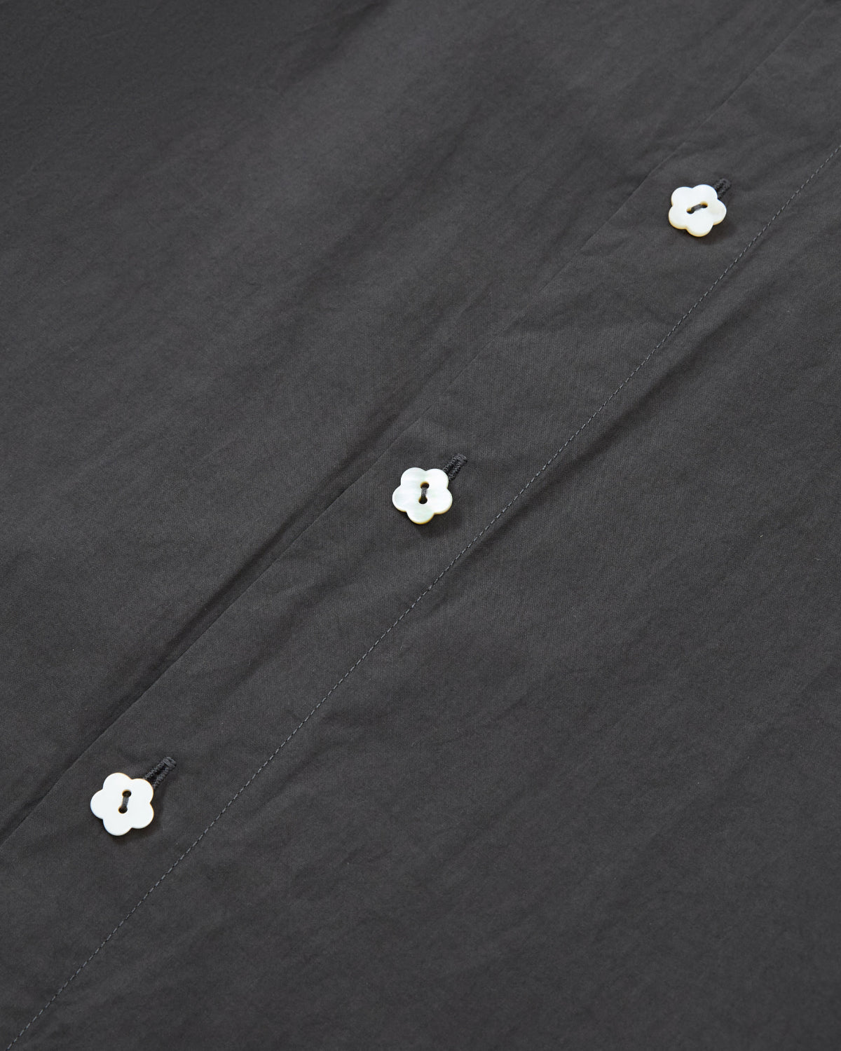 Close-up of the CHARCOAL SUNBAE TIE SHIRT by Sundae School, featuring black fabric adorned with four small, white flower-shaped buttons arranged in a vertical line.