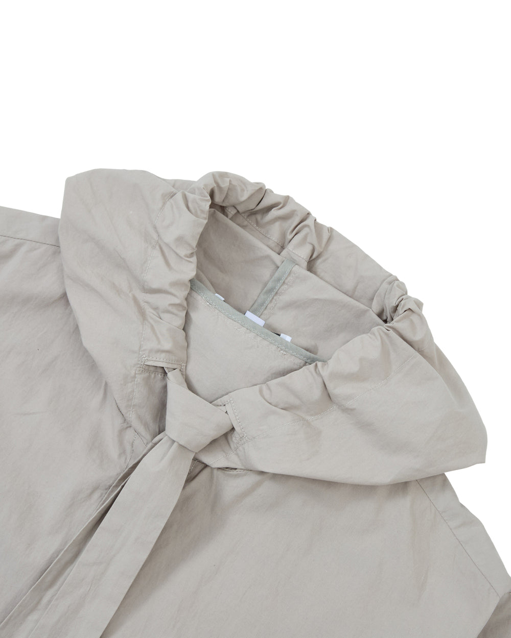 A close-up of the GREY SUNBAE TIE SHIRT by Sundae School, featuring a light beige fabric with a gathered, elastic hood and a partially visible collar, laid flat on a white surface.