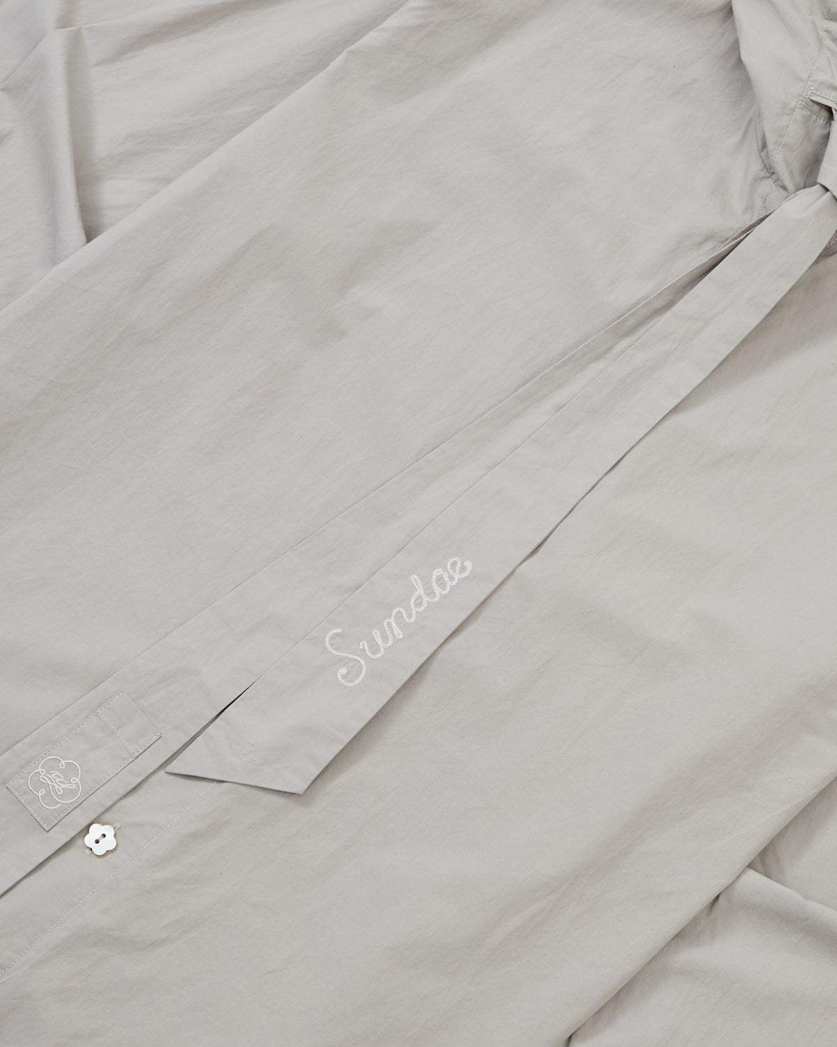 The GREY SUNBAE TIE SHIRT by Sundae School is laid flat, displaying its collar and fabric belt featuring “Sundoo” embroidered in white script, along with a small white embroidered graphic near the cuff.