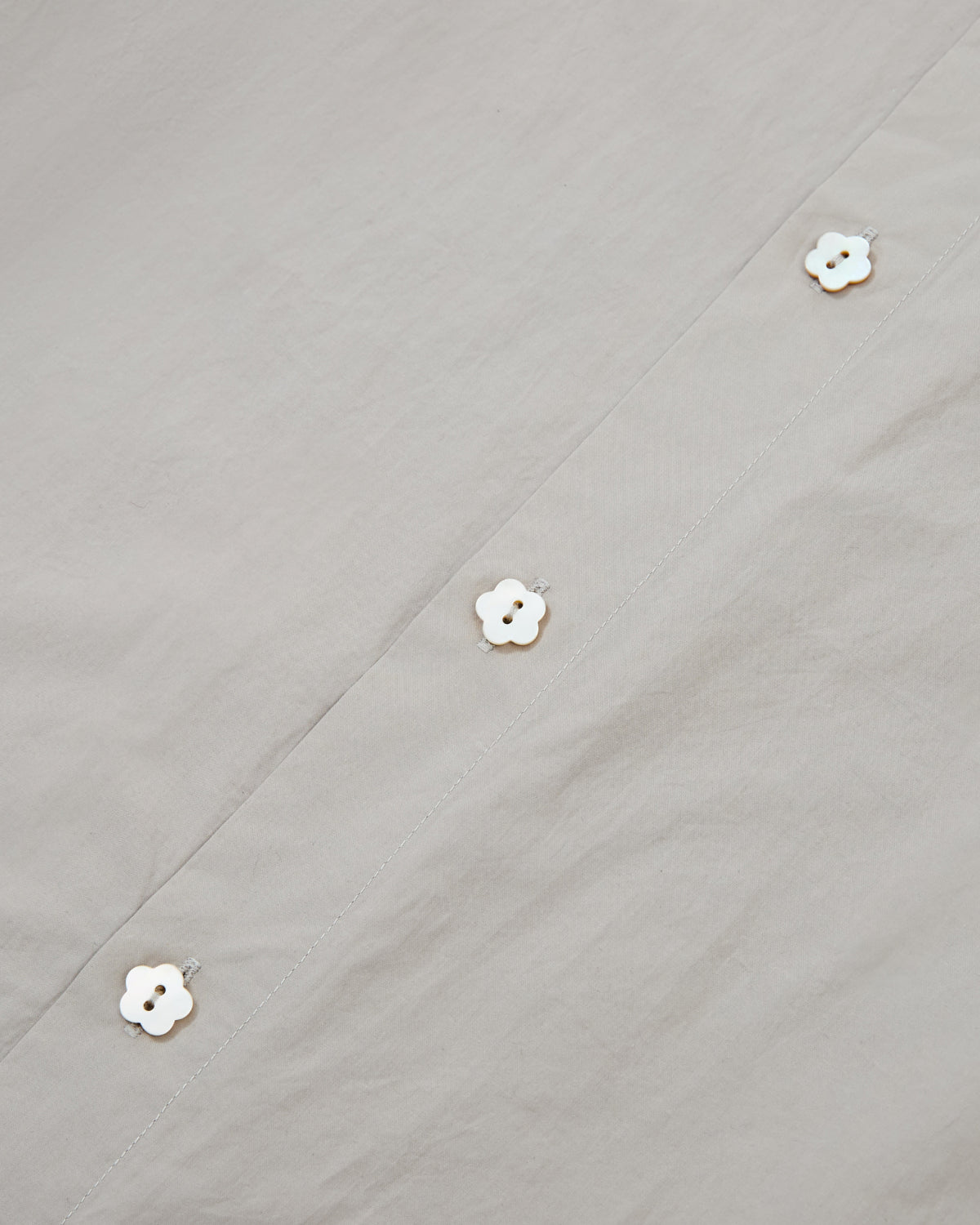 Close-up of the GREY SUNBAE TIE SHIRT by Sundae School, featuring light gray fabric adorned with a row of white flower-shaped buttons along the seam for a simple and elegant look.