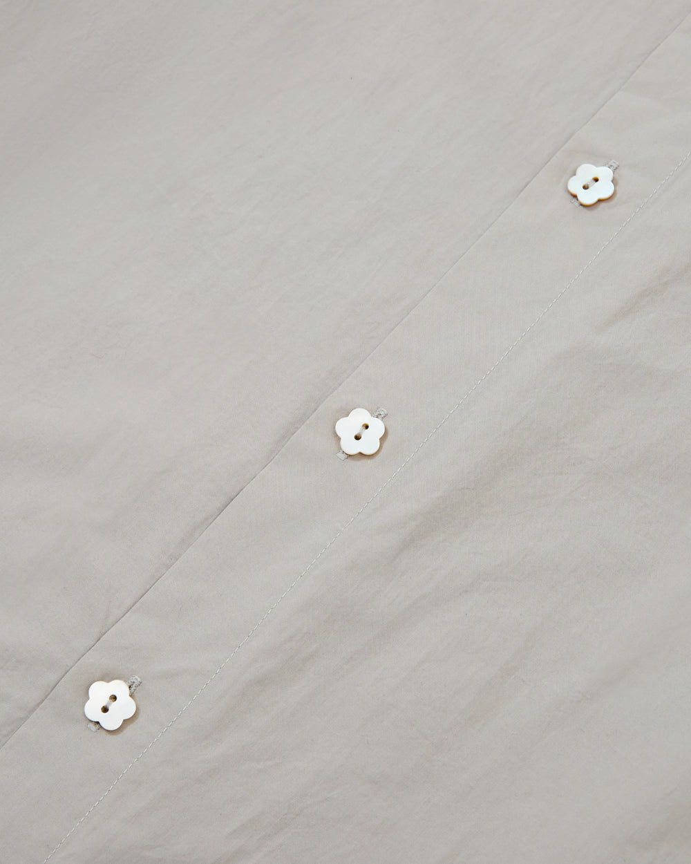 Close-up of the GREY SUNBAE TIE SHIRT by Sundae School, featuring light gray fabric adorned with a row of white flower-shaped buttons along the seam for a simple and elegant look.