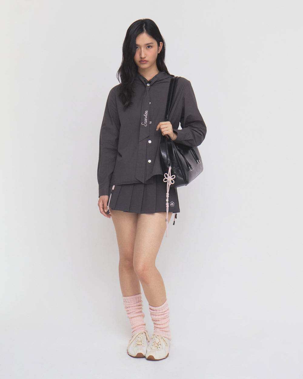 A woman with long dark hair stands against a plain white background, wearing Sundae School’s CHARCOAL SUNBAE TIE SHIRT as a dark button-up jacket, paired with a pleated skirt, pink leg warmers, white sneakers, and carrying a black bag adorned with a pink string accessory.