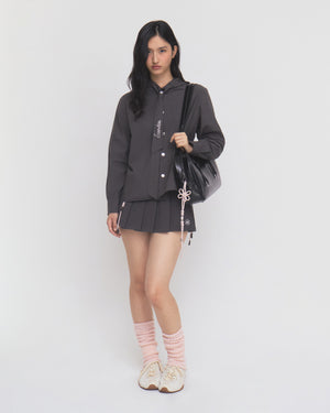 A woman with long dark hair stands against a plain white background, wearing Sundae School’s CHARCOAL SUNBAE TIE SHIRT as a dark button-up jacket, paired with a pleated skirt, pink leg warmers, white sneakers, and carrying a black bag adorned with a pink string accessory.