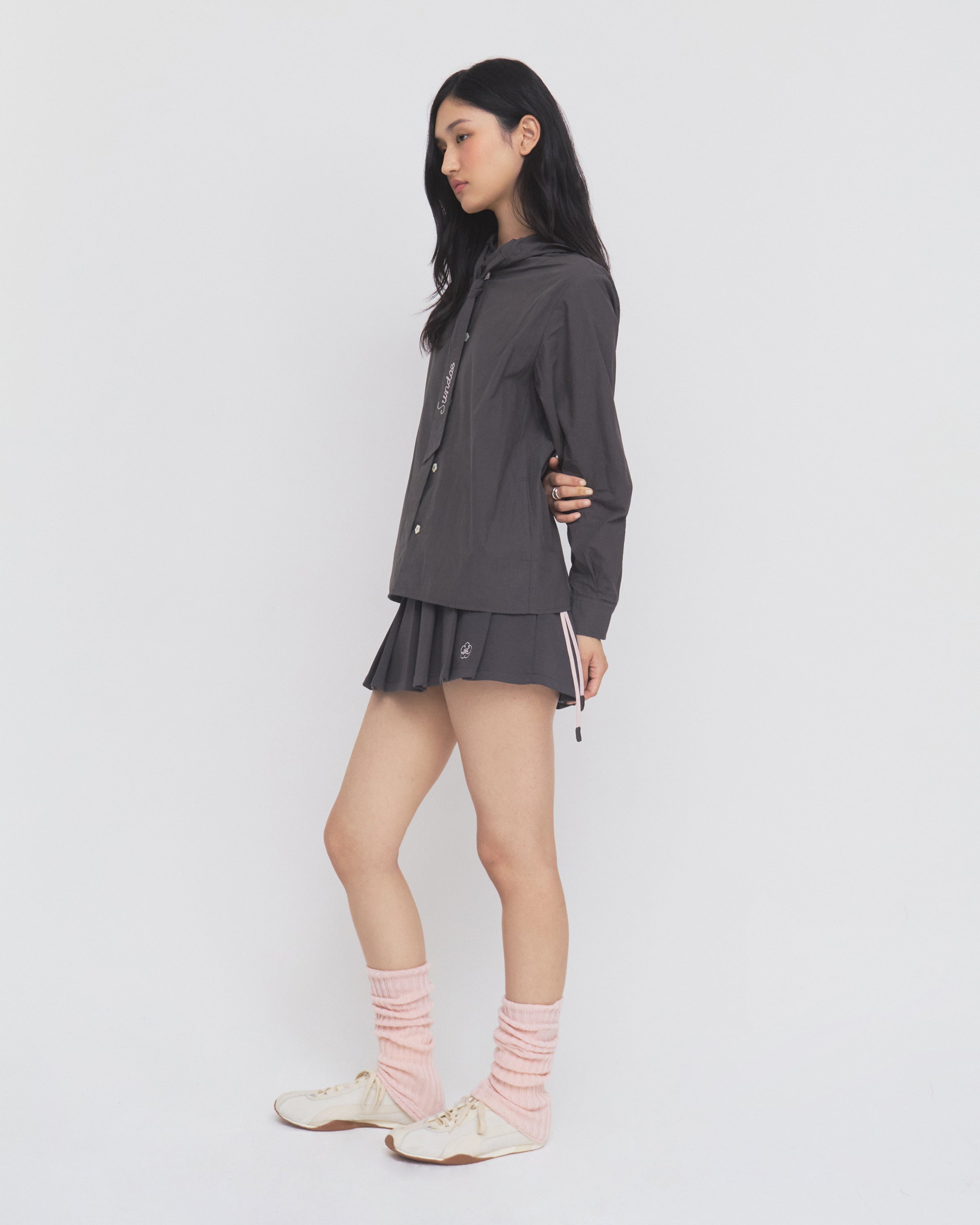 A woman stands sideways against a plain white background, wearing the Sundae School CHARCOAL SUNBAE TIE SHIRT styled with a matching pleated skirt, pink leg warmers, and white sneakers.