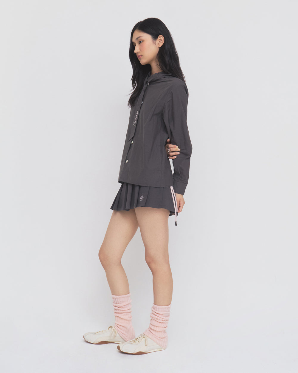 A woman stands sideways against a plain white background, wearing the Sundae School CHARCOAL SUNBAE TIE SHIRT styled with a matching pleated skirt, pink leg warmers, and white sneakers.