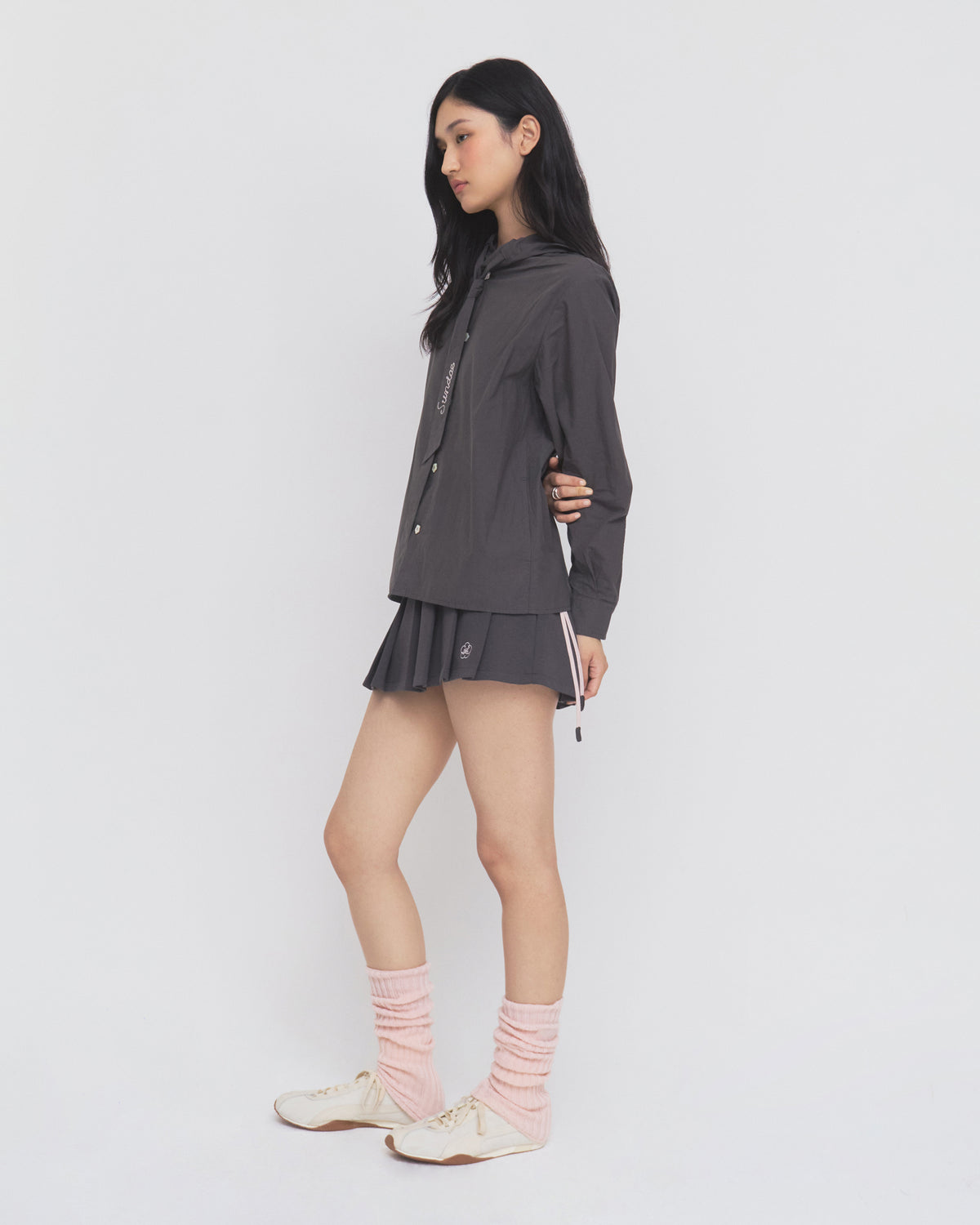 A woman stands sideways against a plain white background, wearing the Sundae School CHARCOAL SUNBAE TIE SHIRT styled with a matching pleated skirt, pink leg warmers, and white sneakers.