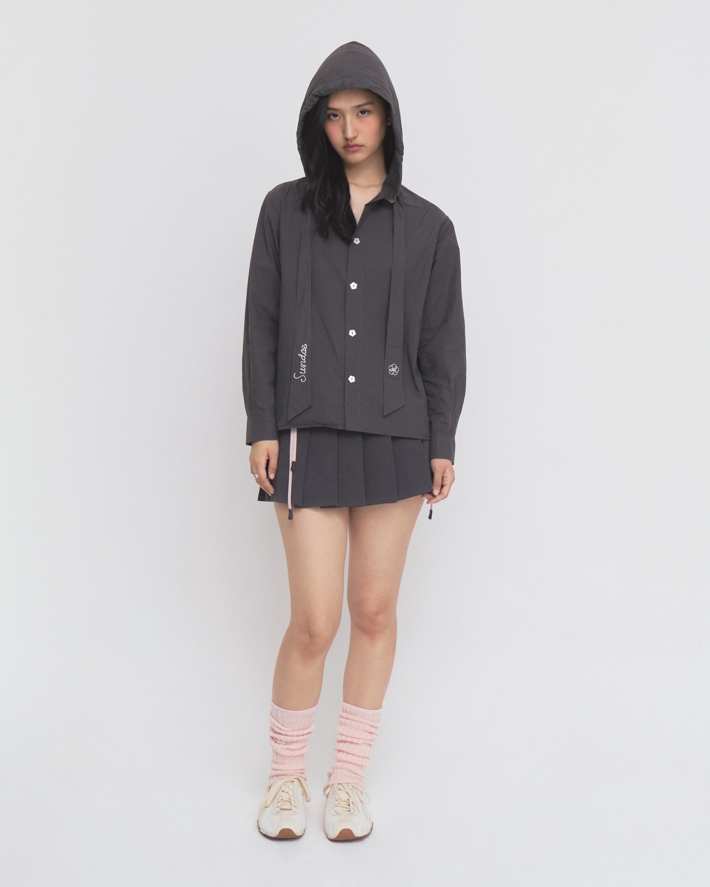 A woman stands against a plain white background wearing the CHARCOAL SUNBAE TIE SHIRT by Sundae School, paired with a matching pleated skirt, white sneakers, and pink scrunched socks. She looks at the camera with a neutral expression, her hands at her sides.